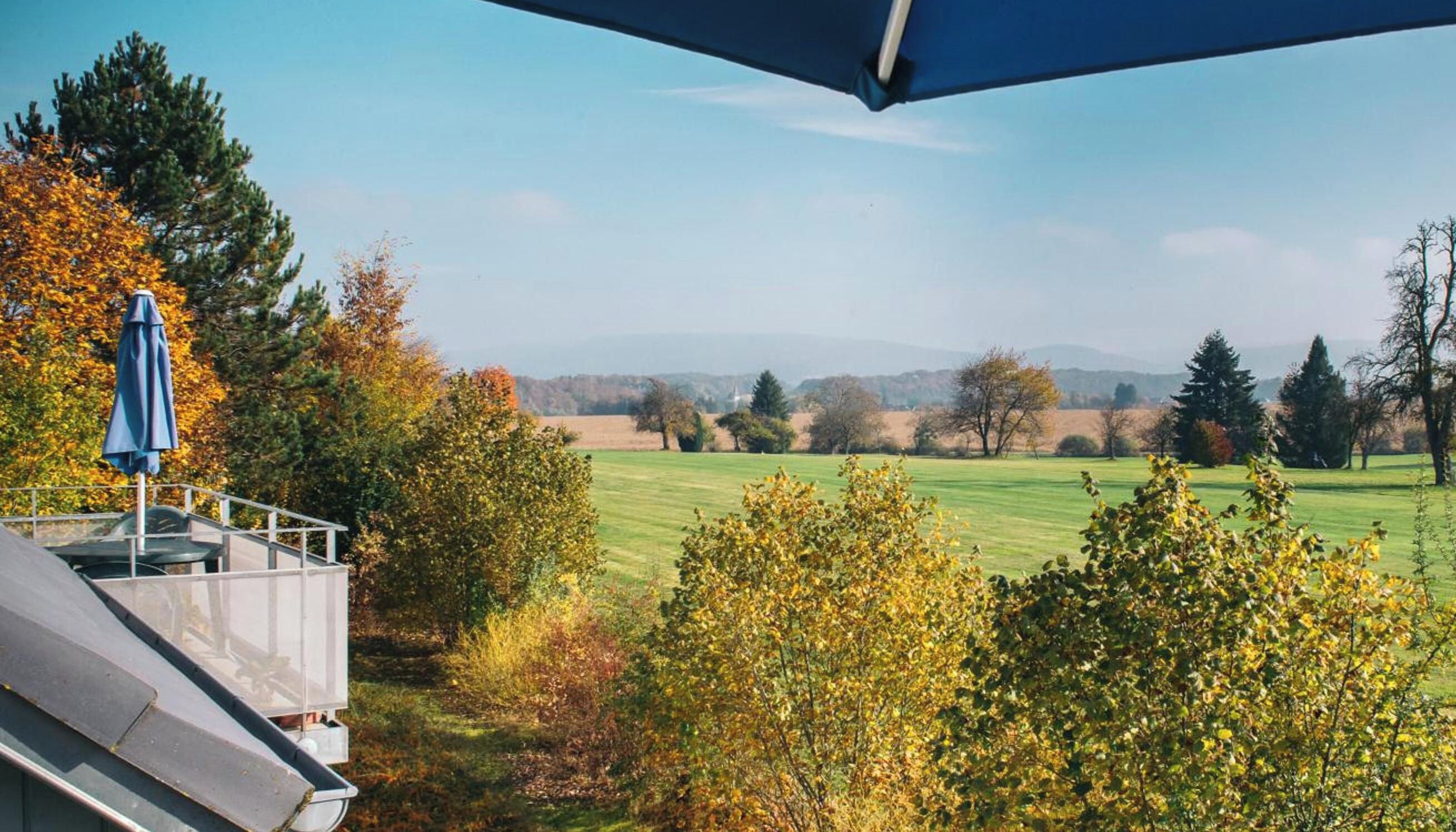 A view from a balcony overlooking a grassy field with trees in autumn colors, a clear sky, and a distant mountain range.