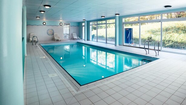 An indoor swimming pool with clear blue water, surrounded by white tiled floors and walls, with a view of greenery outside through large windows. - Residence le Royal Golf & Horse (Mooslargue)