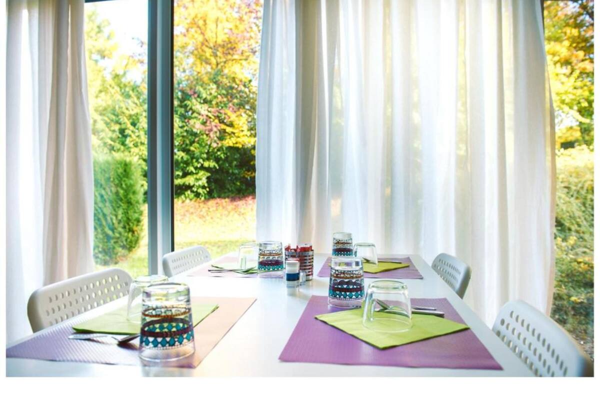 Uma mesa de jantar com potes de vidro coloridos, cadeiras brancas e vista para o verde através de cortinas translúcidas.
