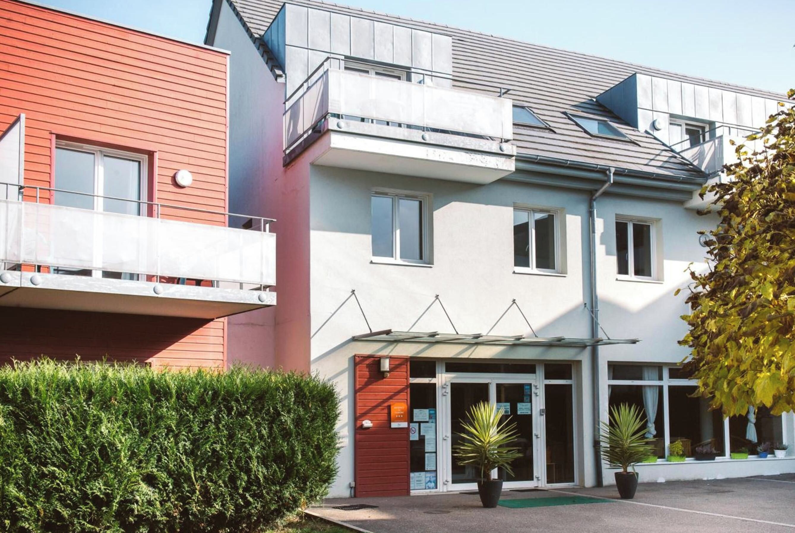 Un edificio residencial de varios pisos con fachada roja y blanca, un acceso cubierto y un pequeño jardín con plantas en macetas.