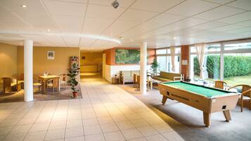 A pool table in a well-lit indoor space with a high ceiling, wooden furniture, and large windows allowing natural light.