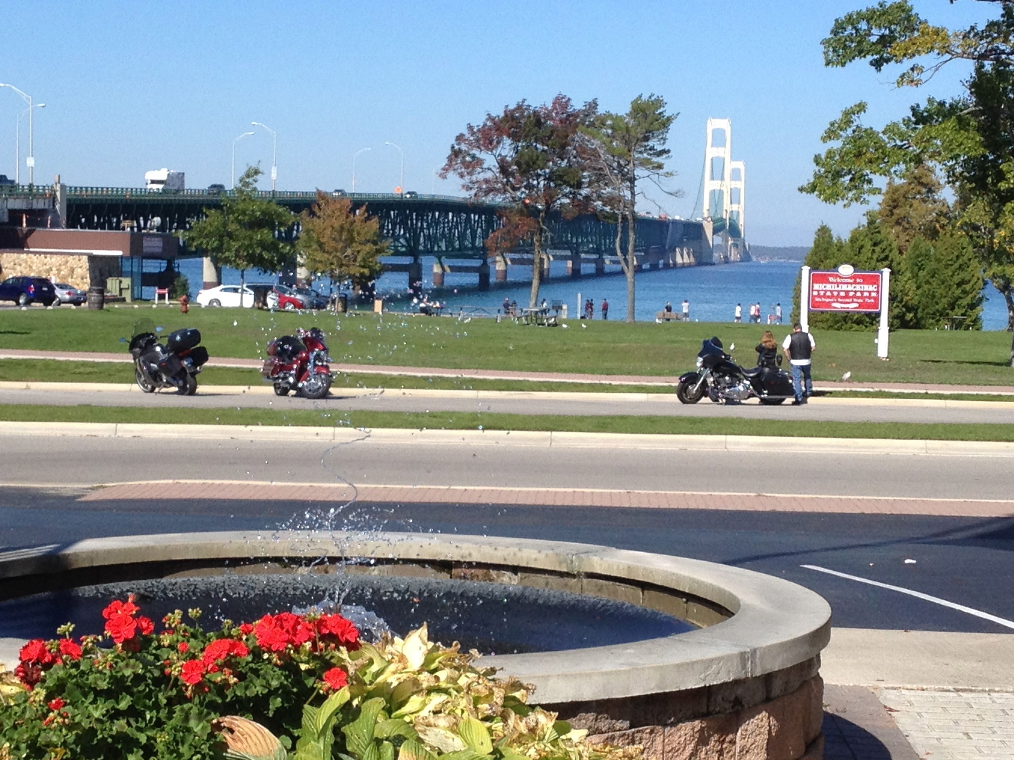 A fountain with water jets, surrounded by red flowers. In the background, there is a bridge over water, a sign for a park, and a few parked motorcycles.