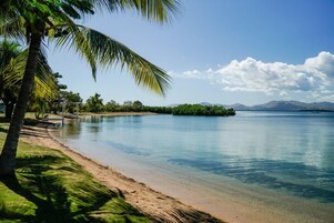 Una playa con agua cristalina, palmeras y montañas a lo lejos.