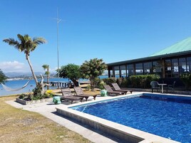 Piscina con sillas de descanso y una hamaca, rodeada de palmeras y vistas al mar.