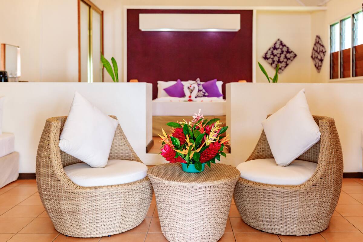 A room with two wicker chairs, a small table with a flower arrangement, a bed with purple pillows, and a mirror on the wall.