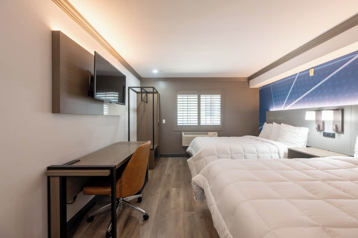 Ein Hotelzimmer mit Schreibtisch, Stuhl, zwei Betten, einem Fernseher und einem Fenster mit Jalousien.