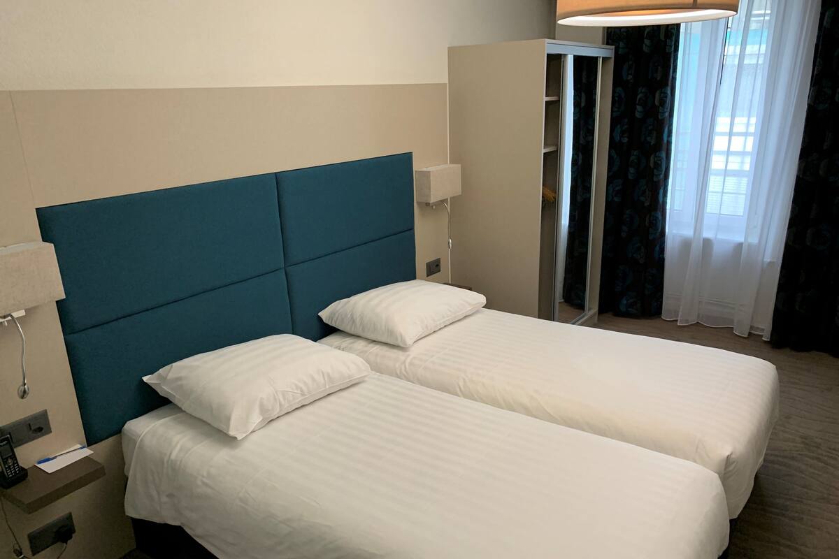 Habitación de hotel con dos camas individuales, cabecera azul, mesita de noche con lámpara y armario.