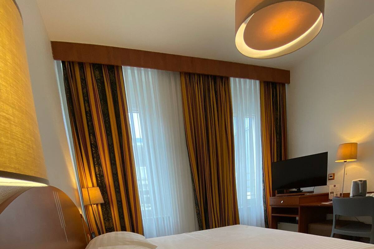 Habitación de hotel con una cama grande, un escritorio con televisión de pantalla plana, una lámpara y cortinas.