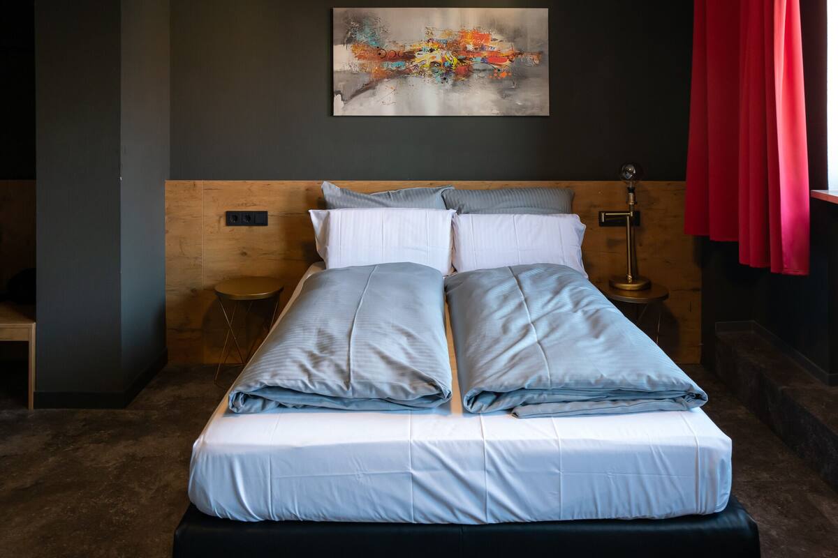 A neatly made bed with a wooden headboard, a bedside table with a lamp, and a red curtain.