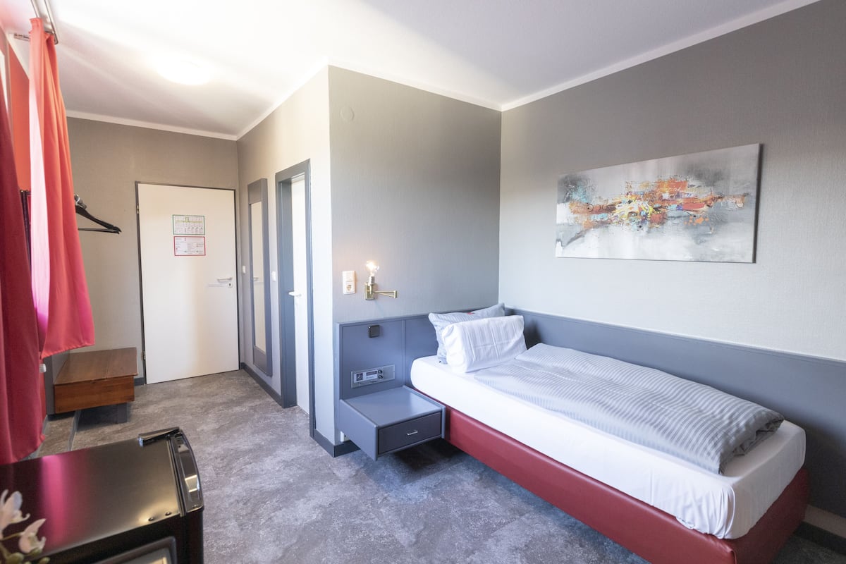 A modern hotel room with a single bed, a small desk, a chair, a wardrobe, and a painting on the wall.