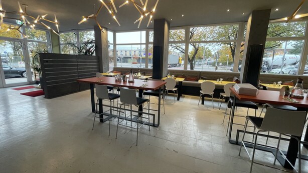 A modern restaurant interior with high ceilings, pendant lighting, and large windows offering views of the street. - Hotel Mecklenheide Hannover (Hannover)