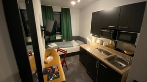 A compact hotel room with a kitchenette, a bed with green curtains, a small dining table with chairs, and a TV mounted on the wall. - Hotel Mecklenheide Hannover (Hannover)
