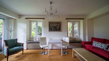 A living room with a green armchair, a red sofa, a white table, and a white chair. There is a chandelier hanging from the ceiling, and a painting on the wall.