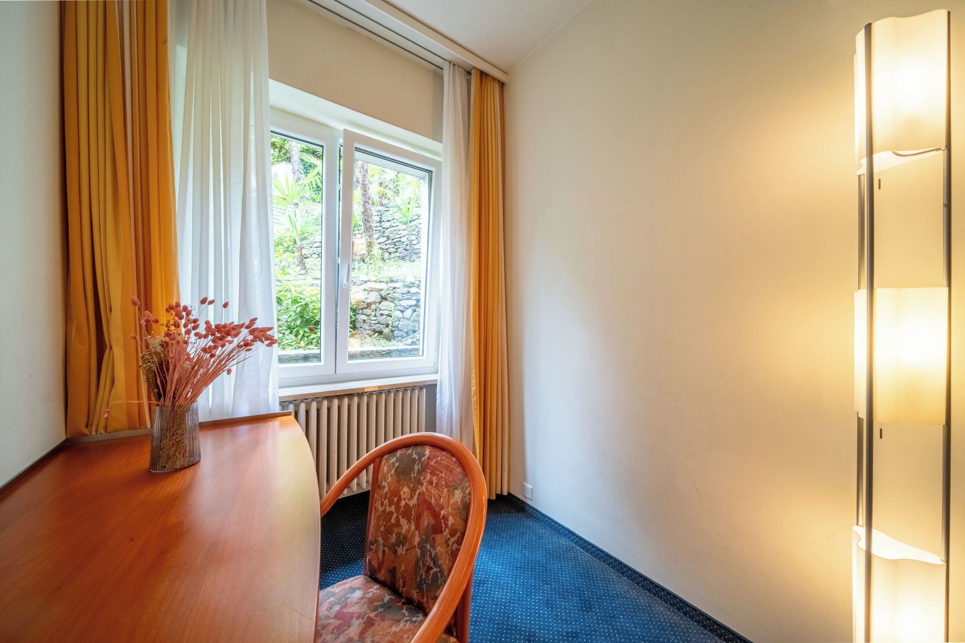 Quarto com mesa de madeira, uma cadeira, janela com cortinas e luminária na parede.