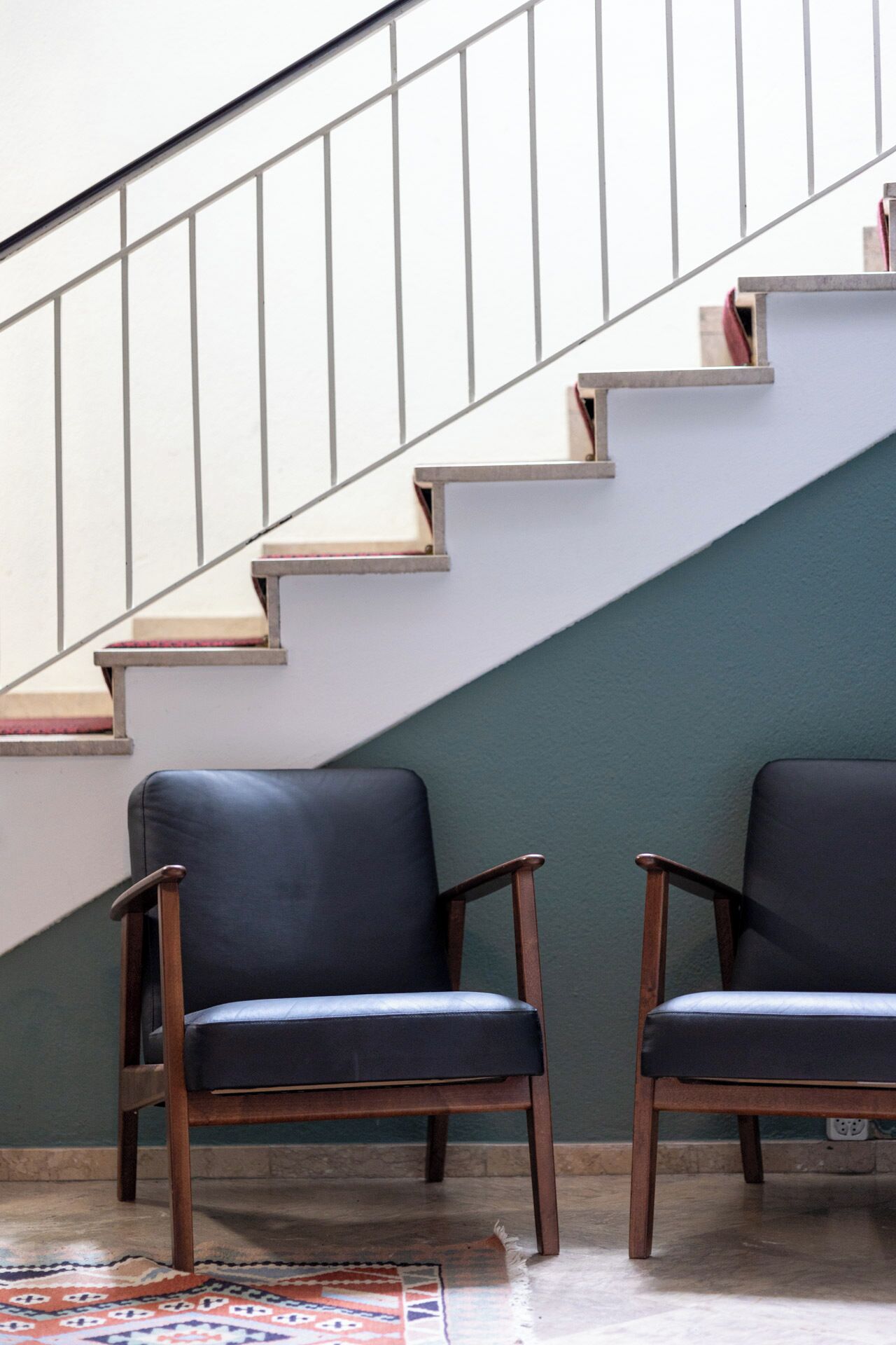 A staircase with a metal railing and a green wall. There are two black leather chairs with wooden legs.