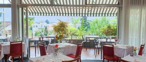 Ein Restaurant mit gedeckten Tischen, einer Pergola mit gestreifter Markise und Blick auf Grünflächen und Gebäude.