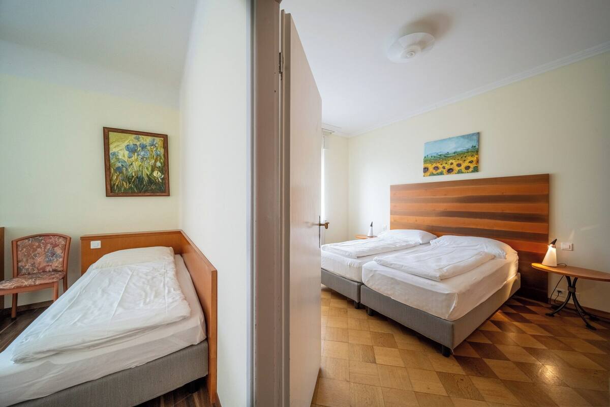 A hotel room with two beds, a wooden headboard, a small table with a lamp, a chair, and two paintings on the walls.