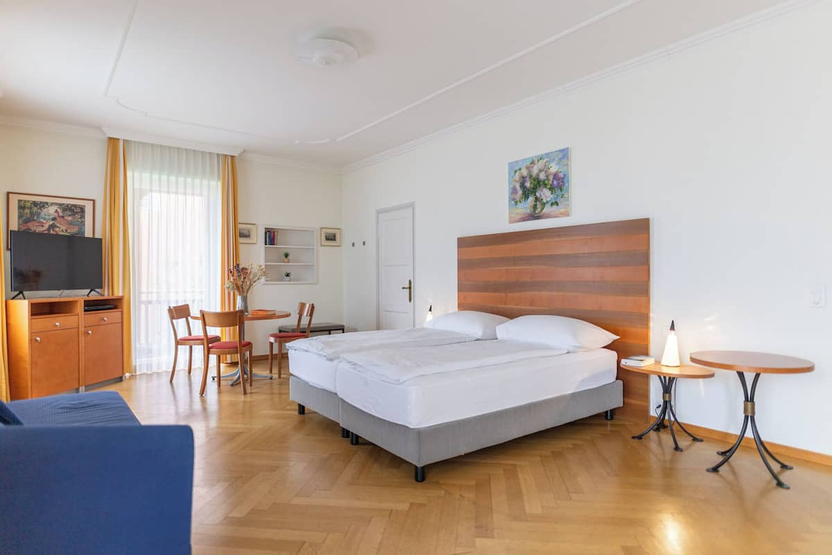 A hotel room with a bed, bedside tables, a desk with chairs, a TV, and a painting on the wall.