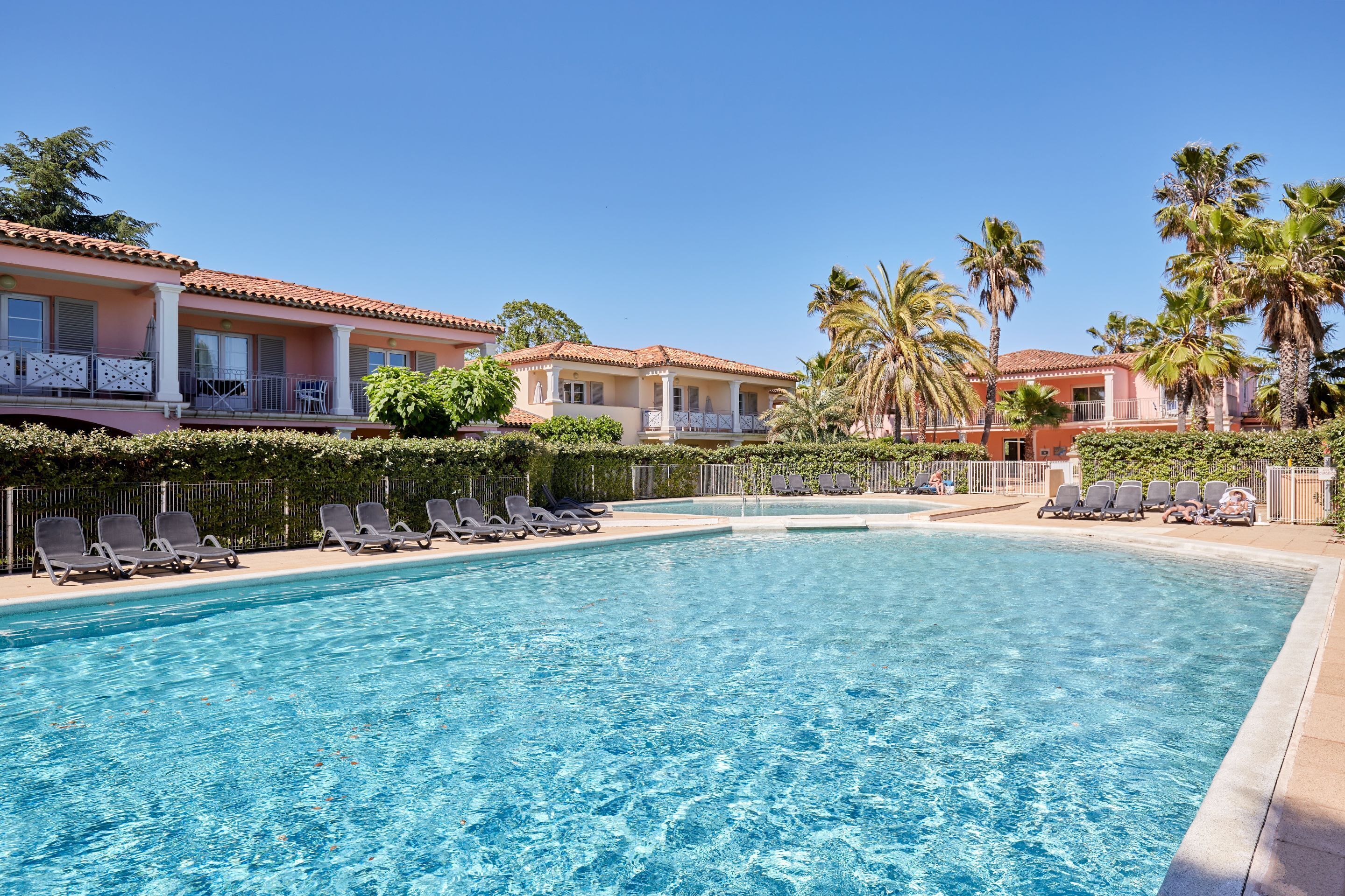 A swimming pool with lounge chairs, palm trees, and a building with a red-tiled roof.