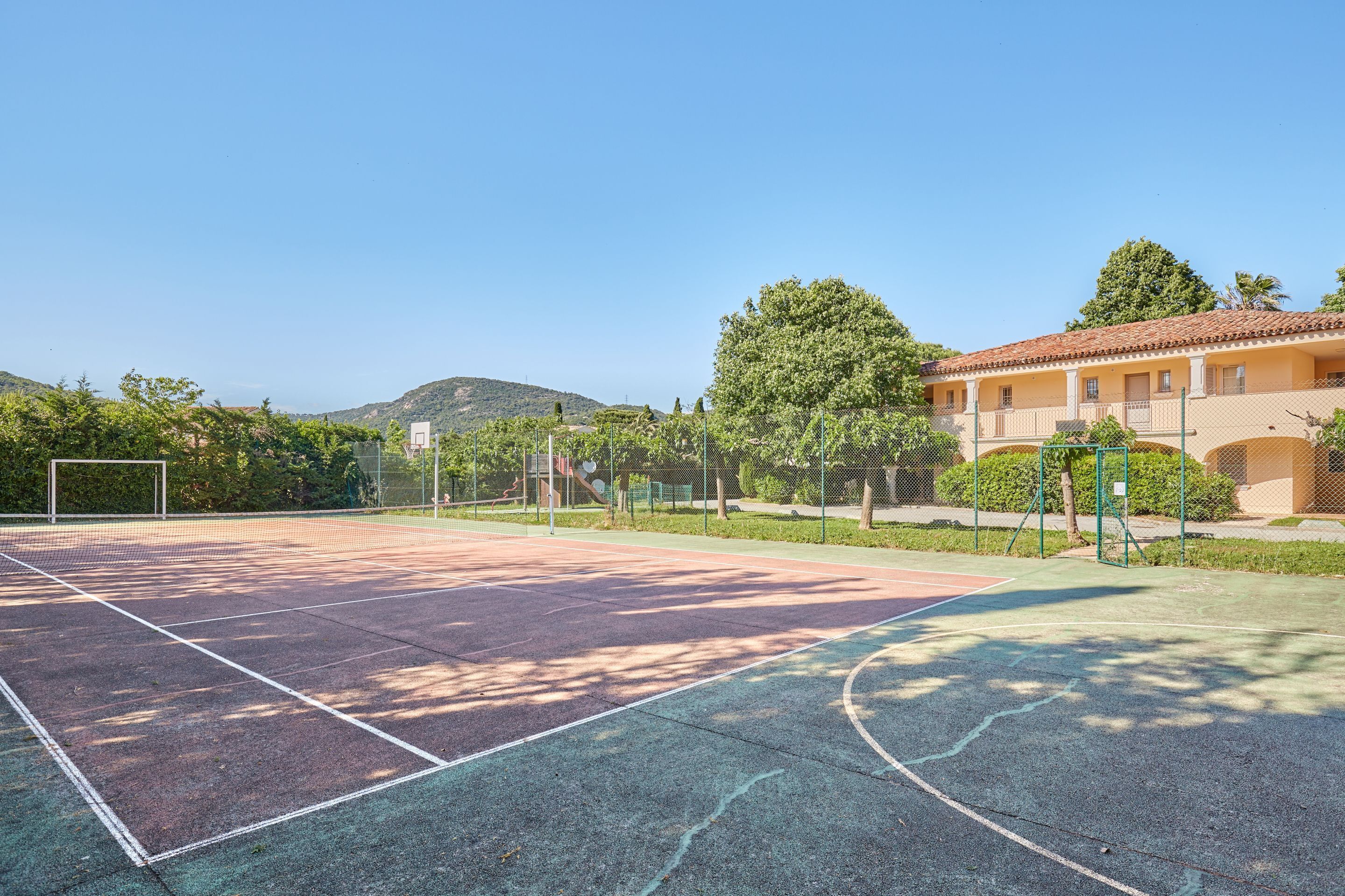 An outdoor tennis court with a net, surrounded by a fence and trees.