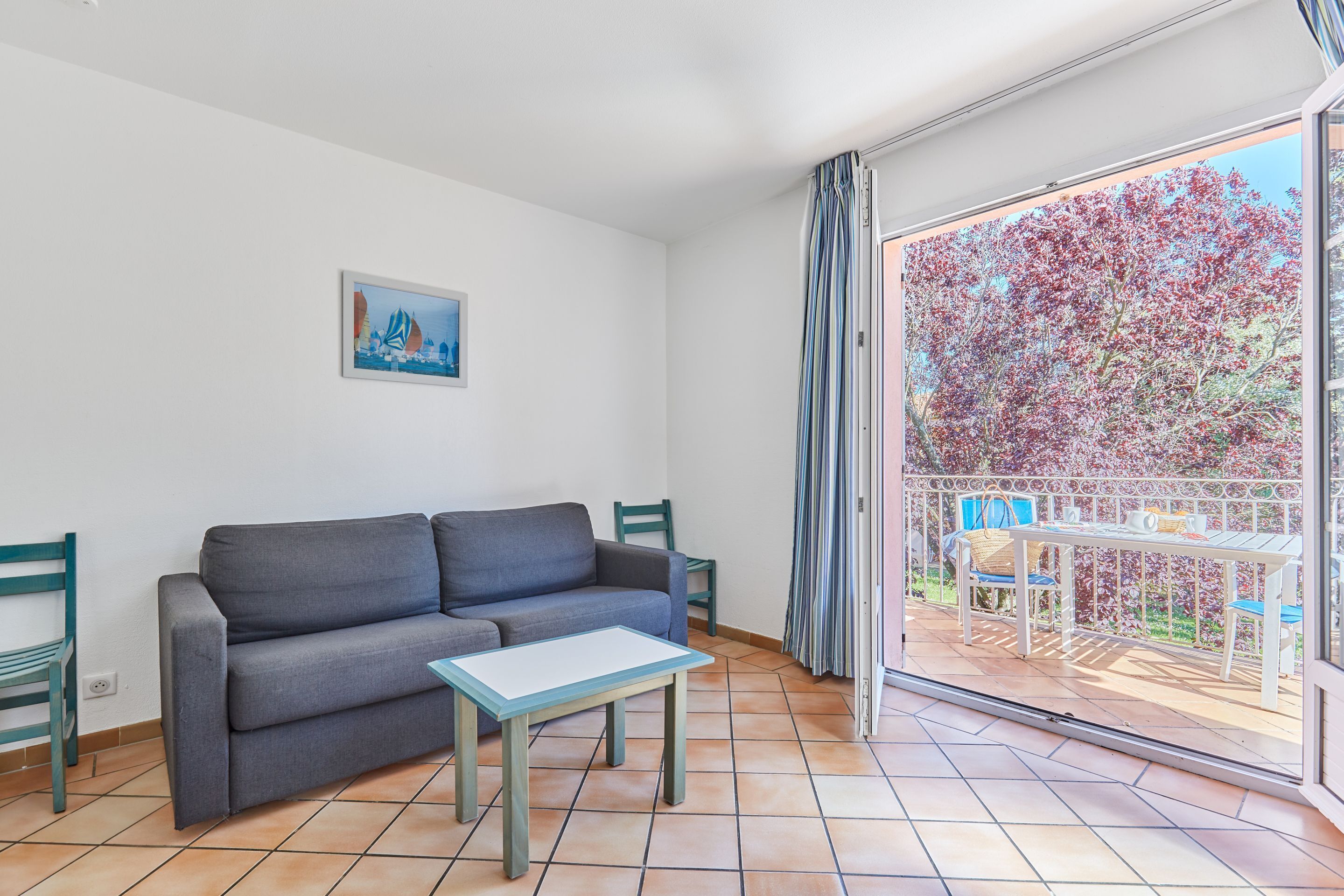 Una sala de estar con un sofá, una mesita y una silla. Cuenta con un balcón con una mesa y sillas.