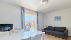 A modern room with a dining table, chairs, a flat-screen TV, a sofa, and a balcony with curtains. - Résidence Odalys La Palmeraie (Grimaud)