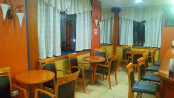 L’intérieur d’un restaurant, avec de nombreuses tables et chaises, de grandes fenêtres ornées de rideaux et un sol carrelé.