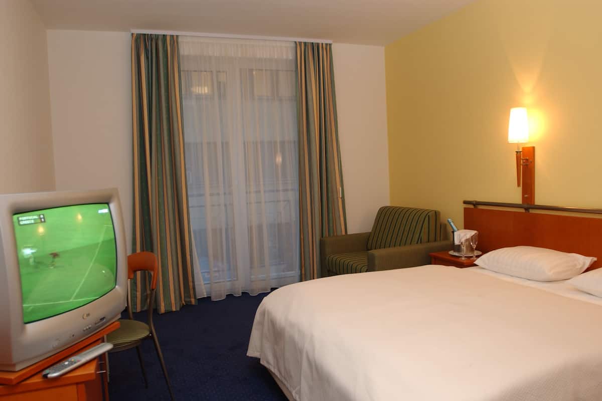 A hotel room with a bed, a television displaying a sports game, a chair, a nightstand with a lamp, and a window with curtains.