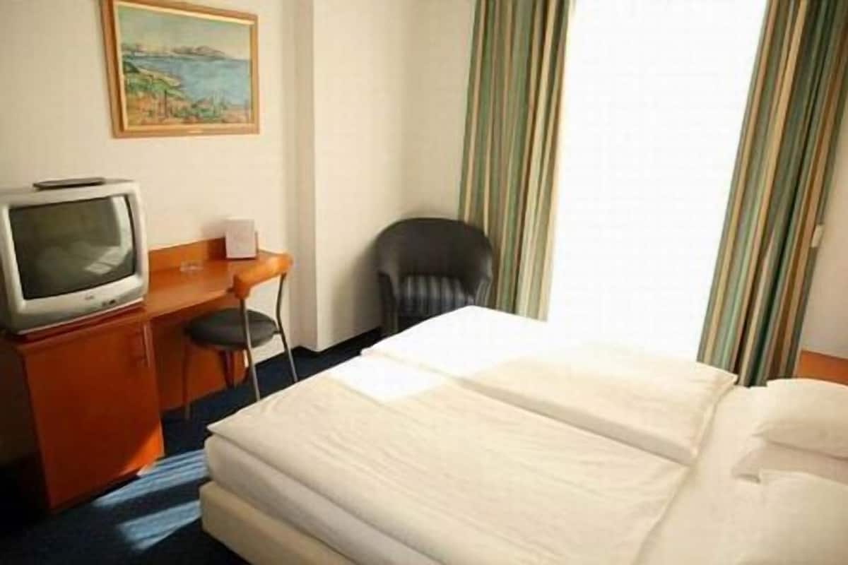A hotel room with a bed, a desk with a television, a chair, and a painting on the wall.