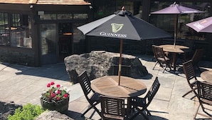 An outdoor seating area with tables and chairs, a stone planter with flowers, and a building with a sloped roof and large windows. - Summit Lodge (Killington)