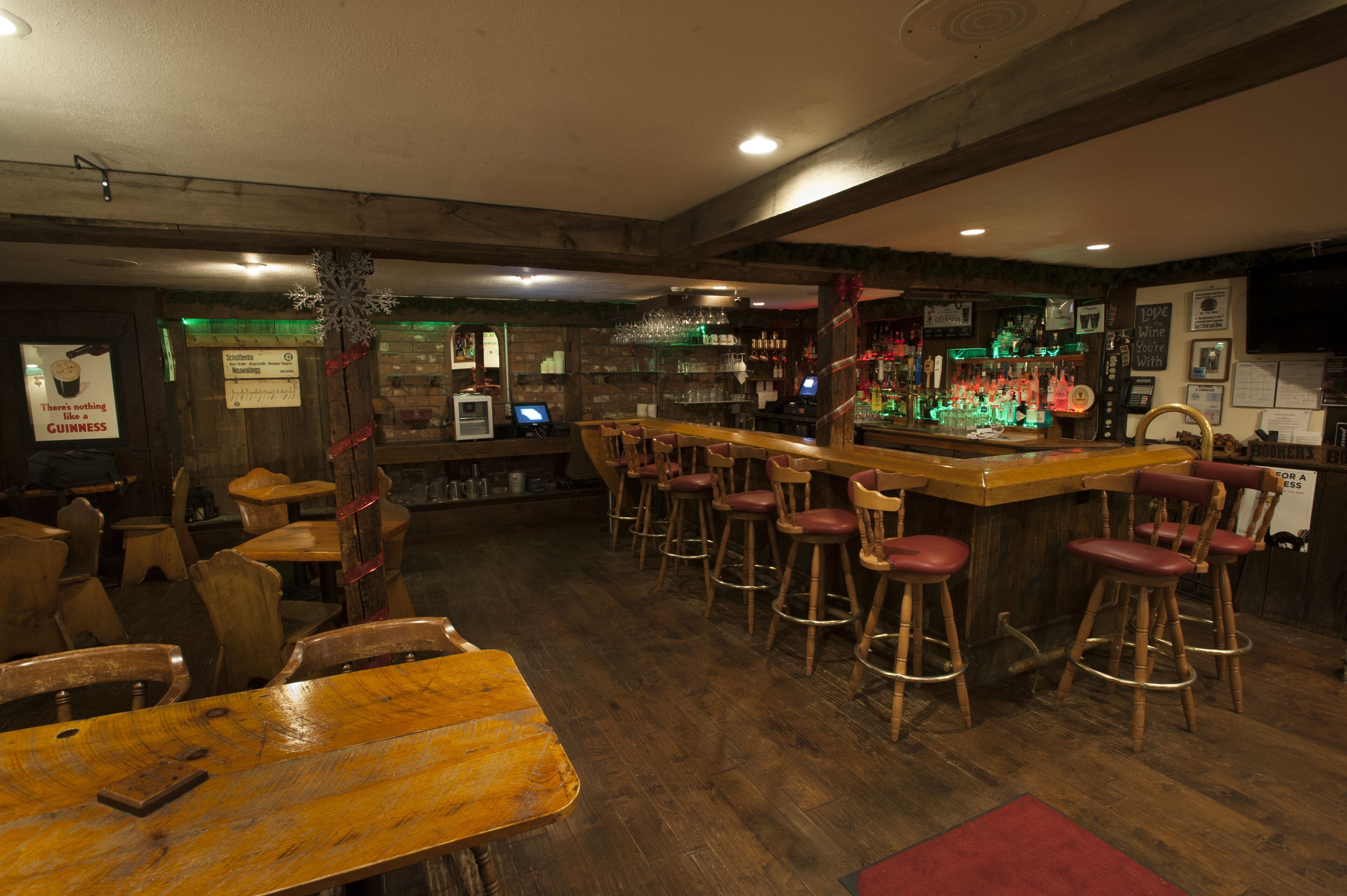 A bar area with wooden tables and stools, a bar counter, and shelves with bottles and glasses.