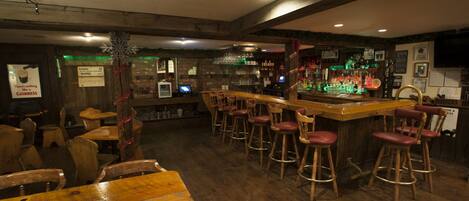 A bar area with wooden tables and stools, a bar counter, and shelves with bottles and glasses.
