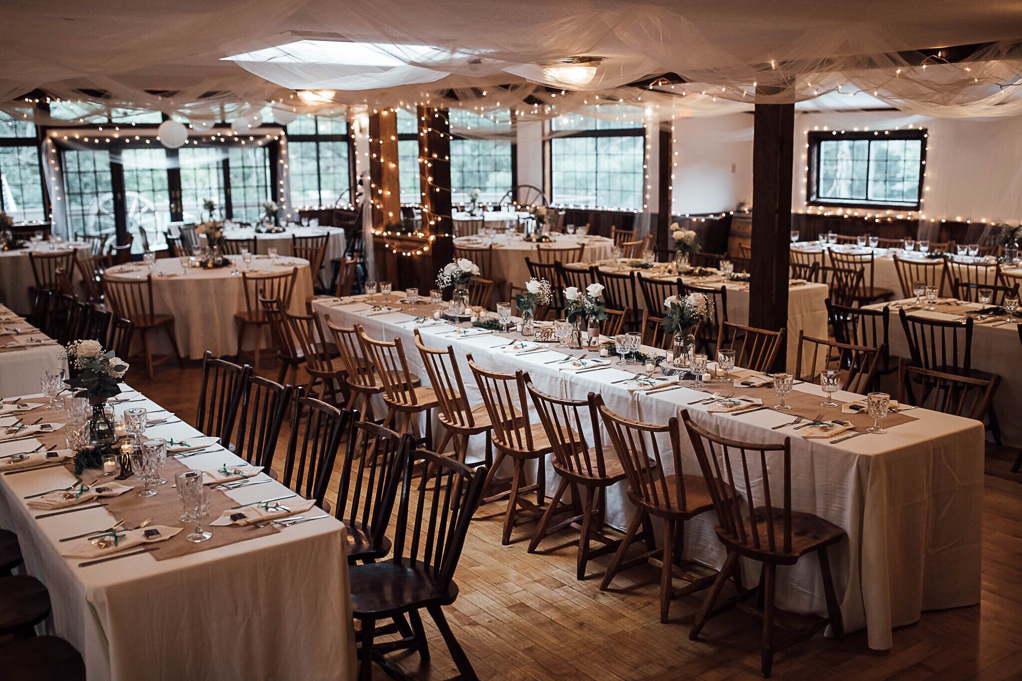Un salón de banquetes con varias mesas redondas, cada una con mantel blanco, sillas de madera y cristalería.