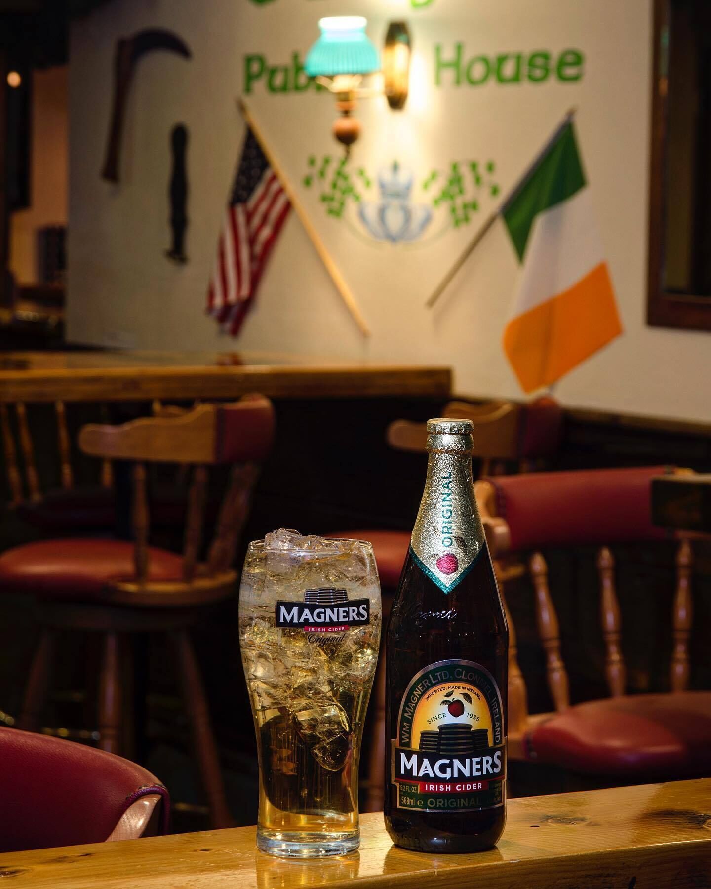 A glass of MAGNERS Irish cider and a bottle of MAGNERS Irish cider on a wooden table in a pub with a sign reading "Pub House".