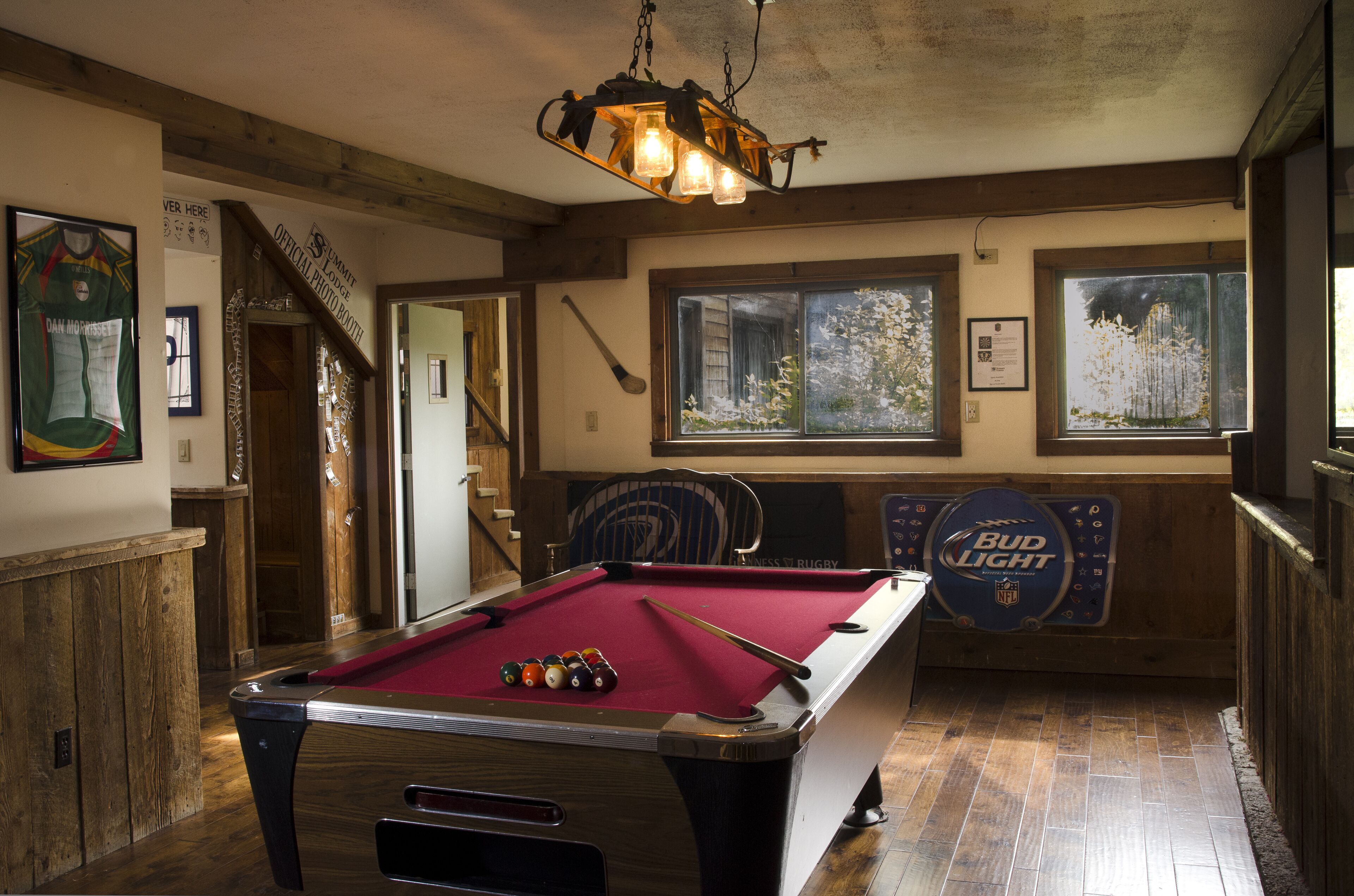 A pool table with balls, a pool cue, and a Bud Light sign in a room with wooden floors and a staircase.