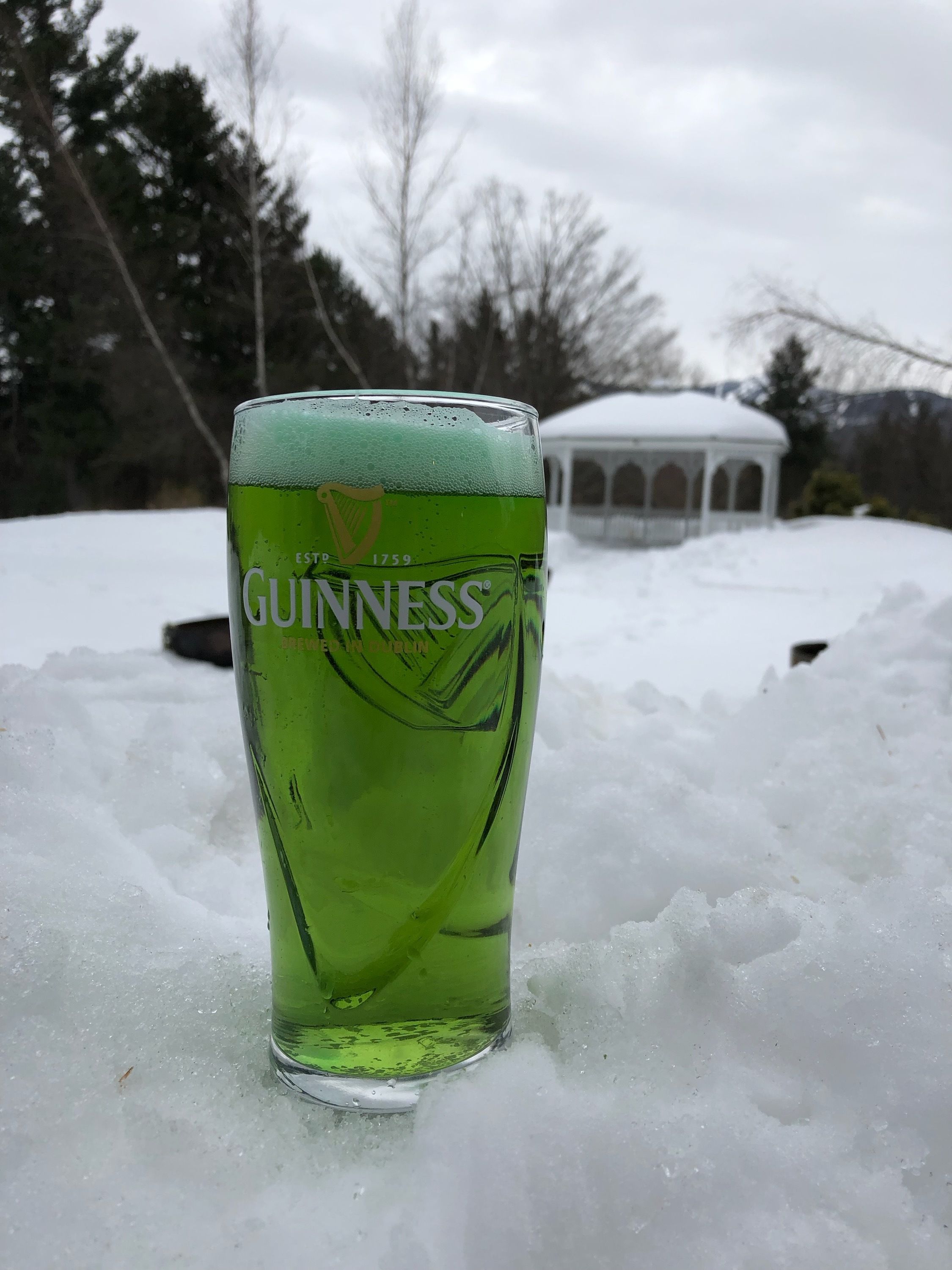 A glass of Guinness beer on a snowy surface with a gazebo in the background.
