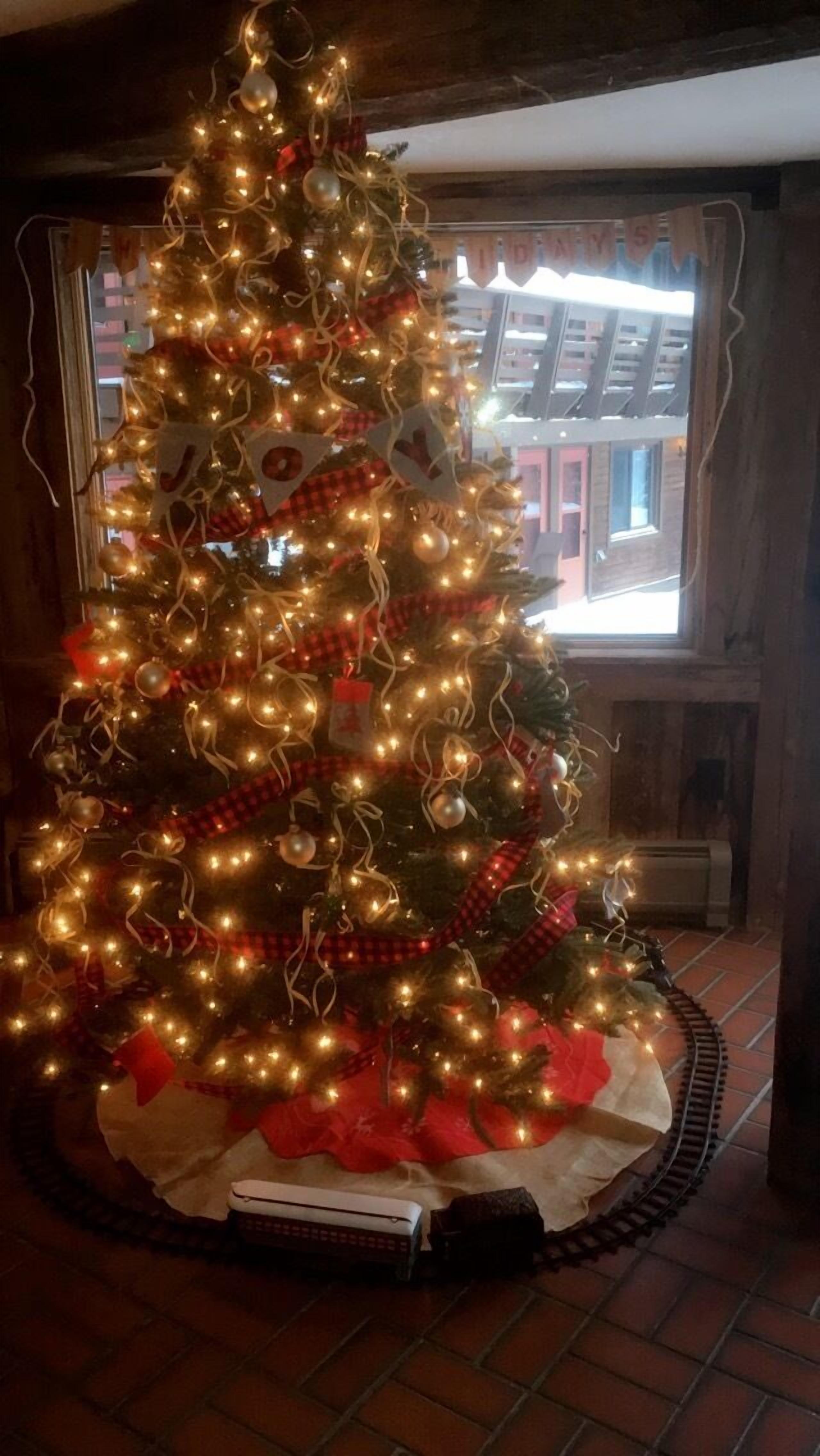 A decorated Christmas tree with lights and ornaments, placed in a room with a brick floor and a window with a view of buildings outside.