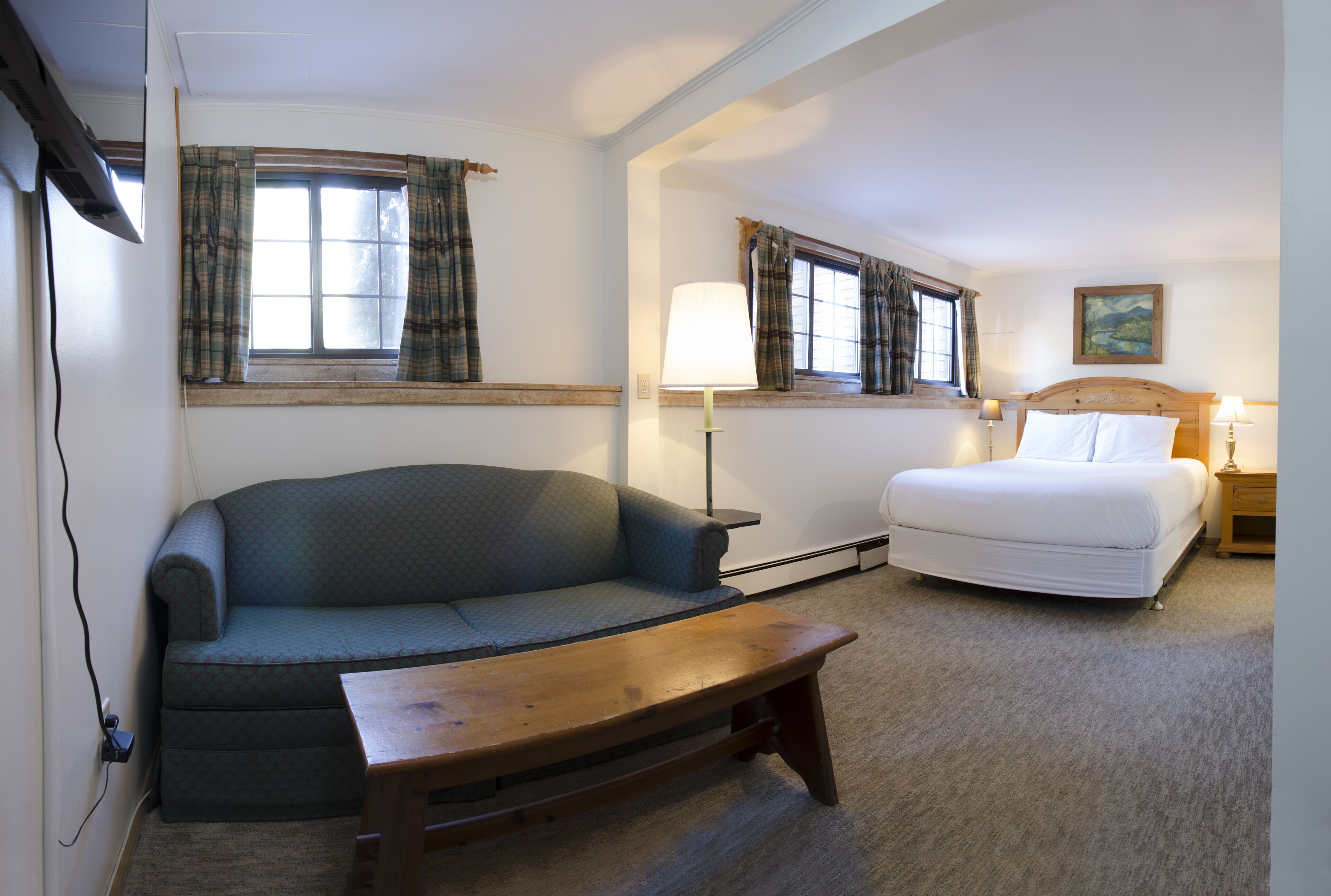 A room with a sofa, a bed, a wooden table, and two windows with curtains.
