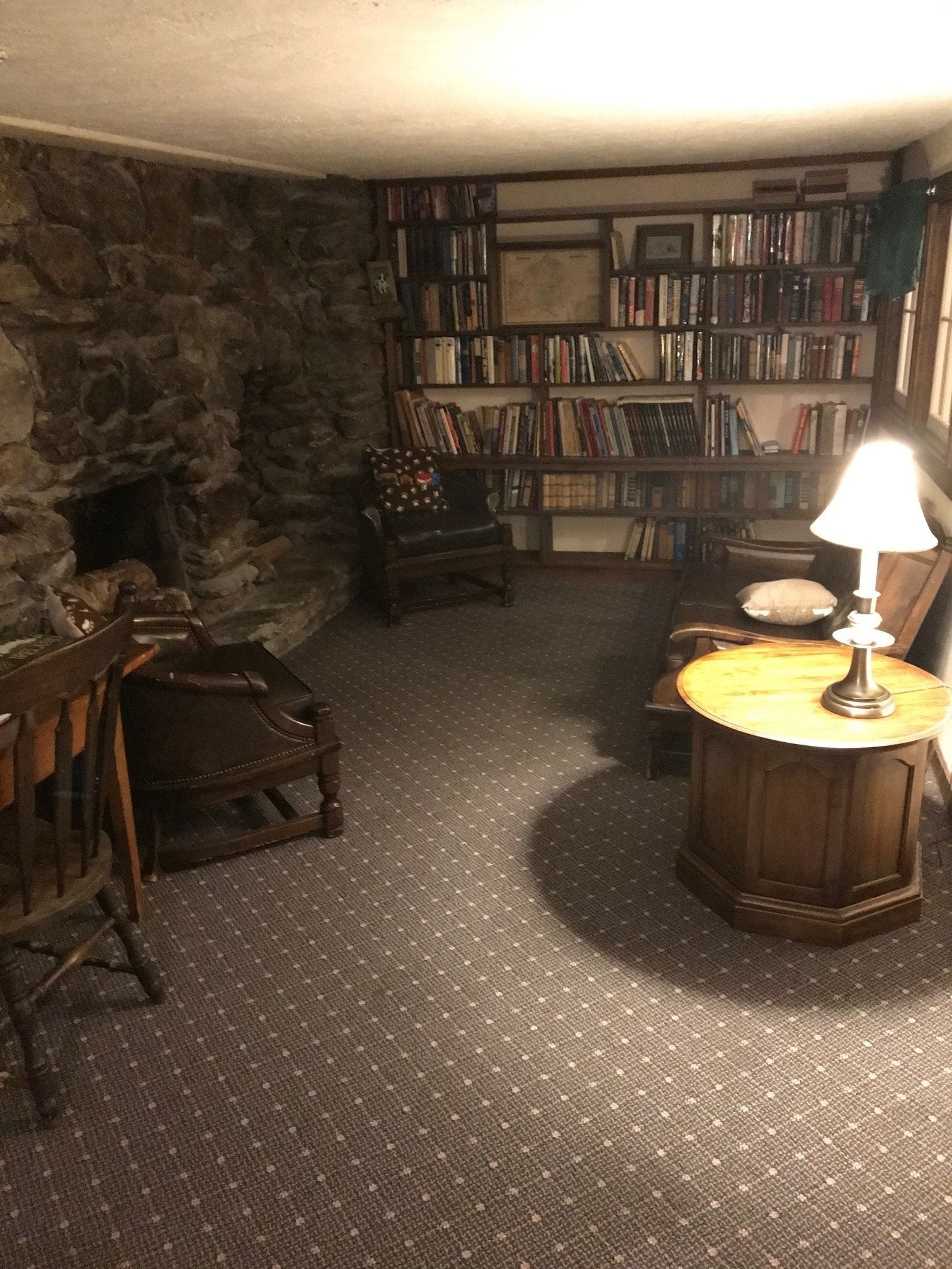 A cozy room with a stone wall, a fireplace, a bookshelf, a wooden chair, a round table with a lamp, and a patterned carpet.