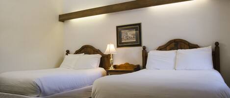 A hotel room with two single beds, wooden headboards, a nightstand with a lamp, and a framed picture on the wall.