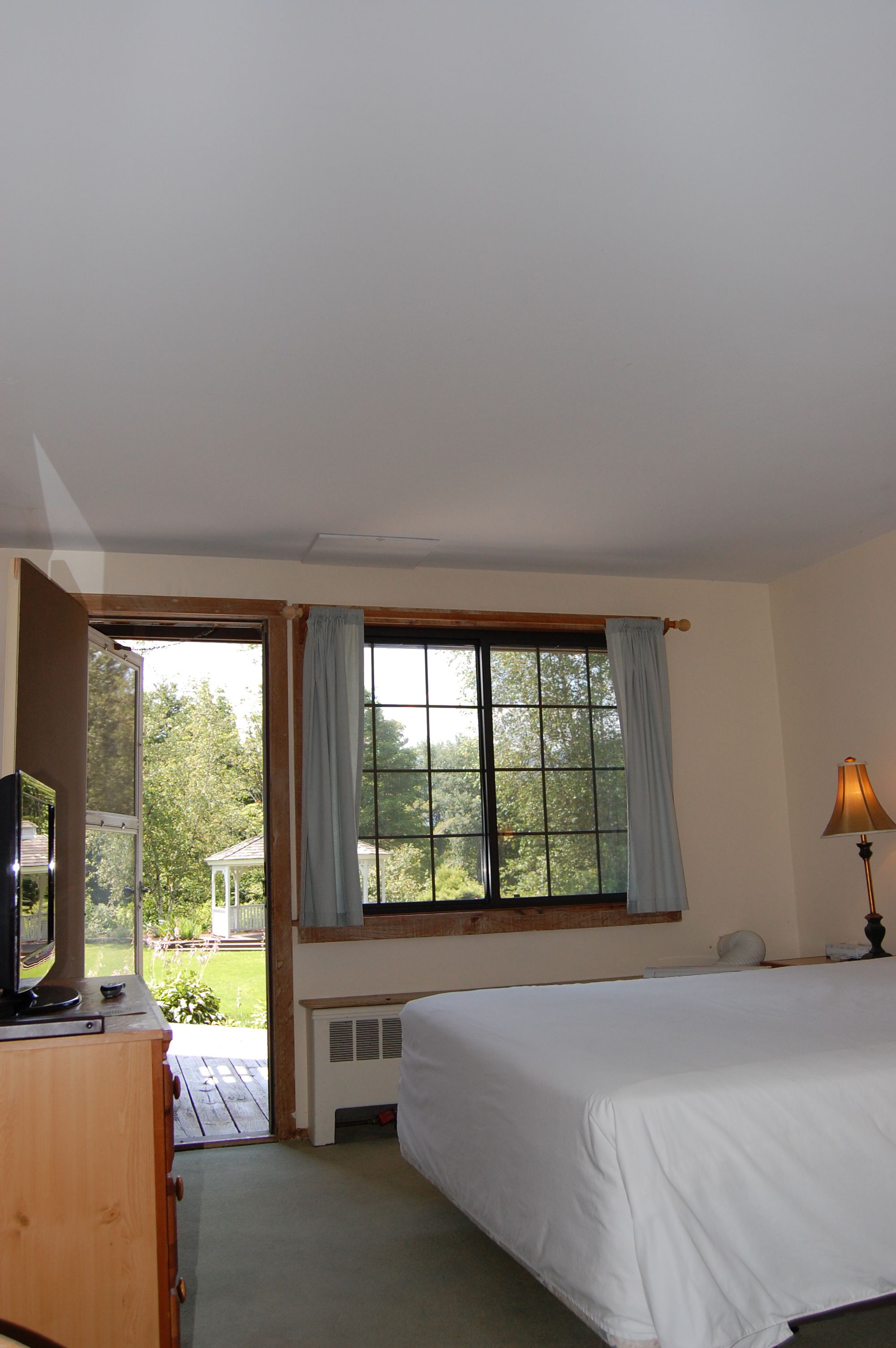 Un dormitorio con una cama grande, un televisor sobre una cómoda de madera, una ventana con cortinas y una lámpara.