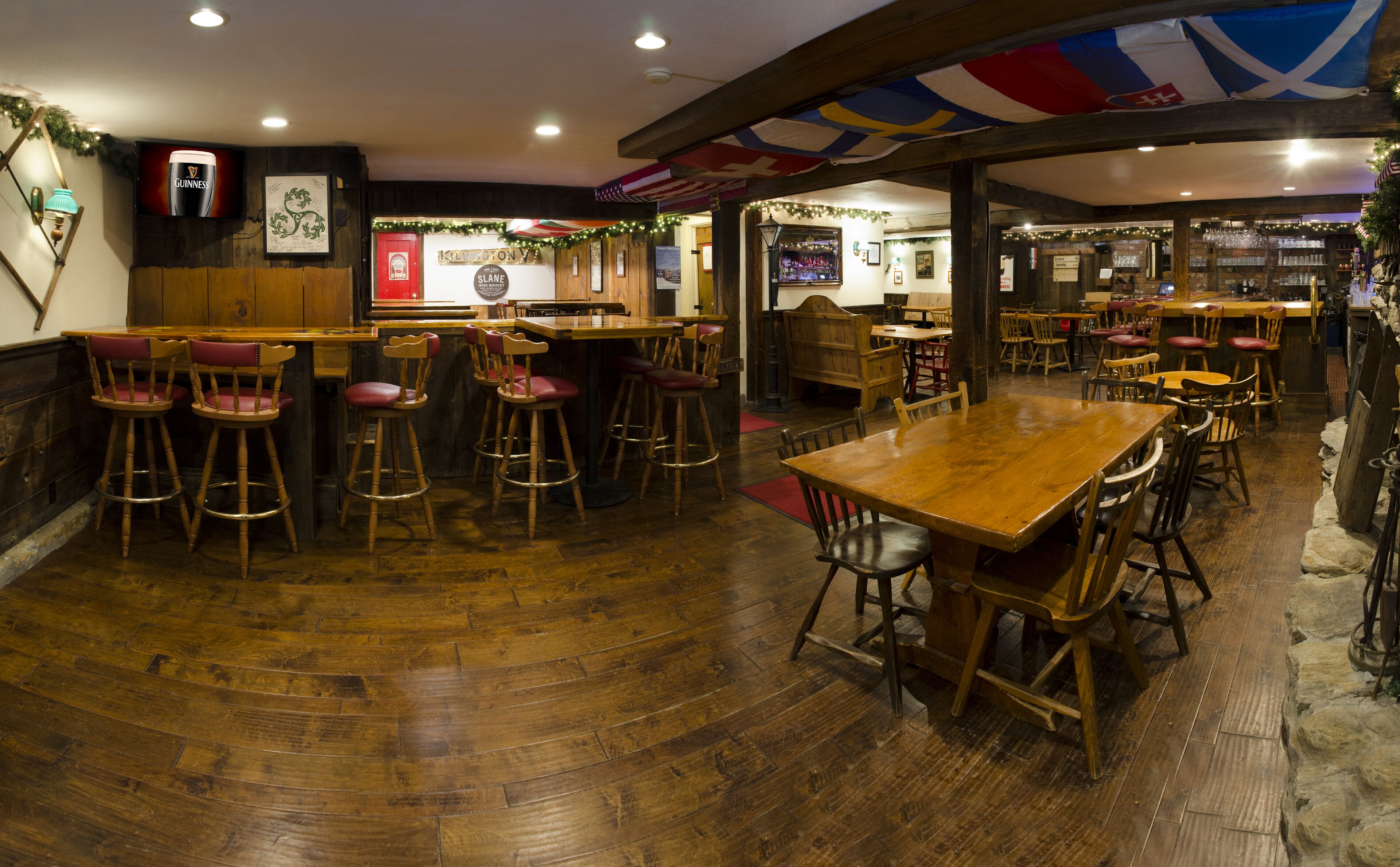 Un espace bar avec des meubles en bois, une grande table en bois et des tabourets. Des drapeaux sont suspendus au plafond et une enseigne indique « KILPATRON SLANE ».