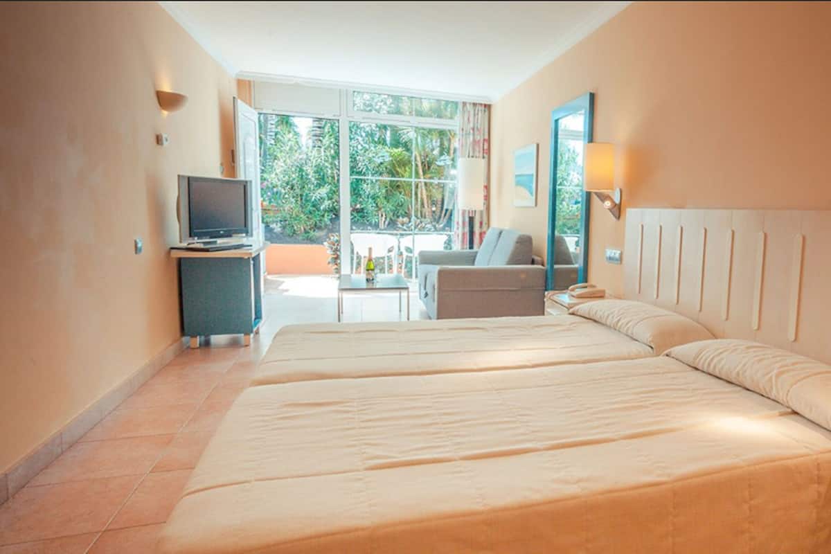 A hotel room with two beds, a television, a desk, and a balcony with a view of greenery.