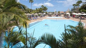 Una piscina rodeada de sillas de descanso y sombrillas, con palmeras y un edificio a lo lejos.