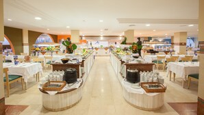 Montaje de bufé con varias estaciones de comida, mesas y sillas dispuestas en un salón amplio.