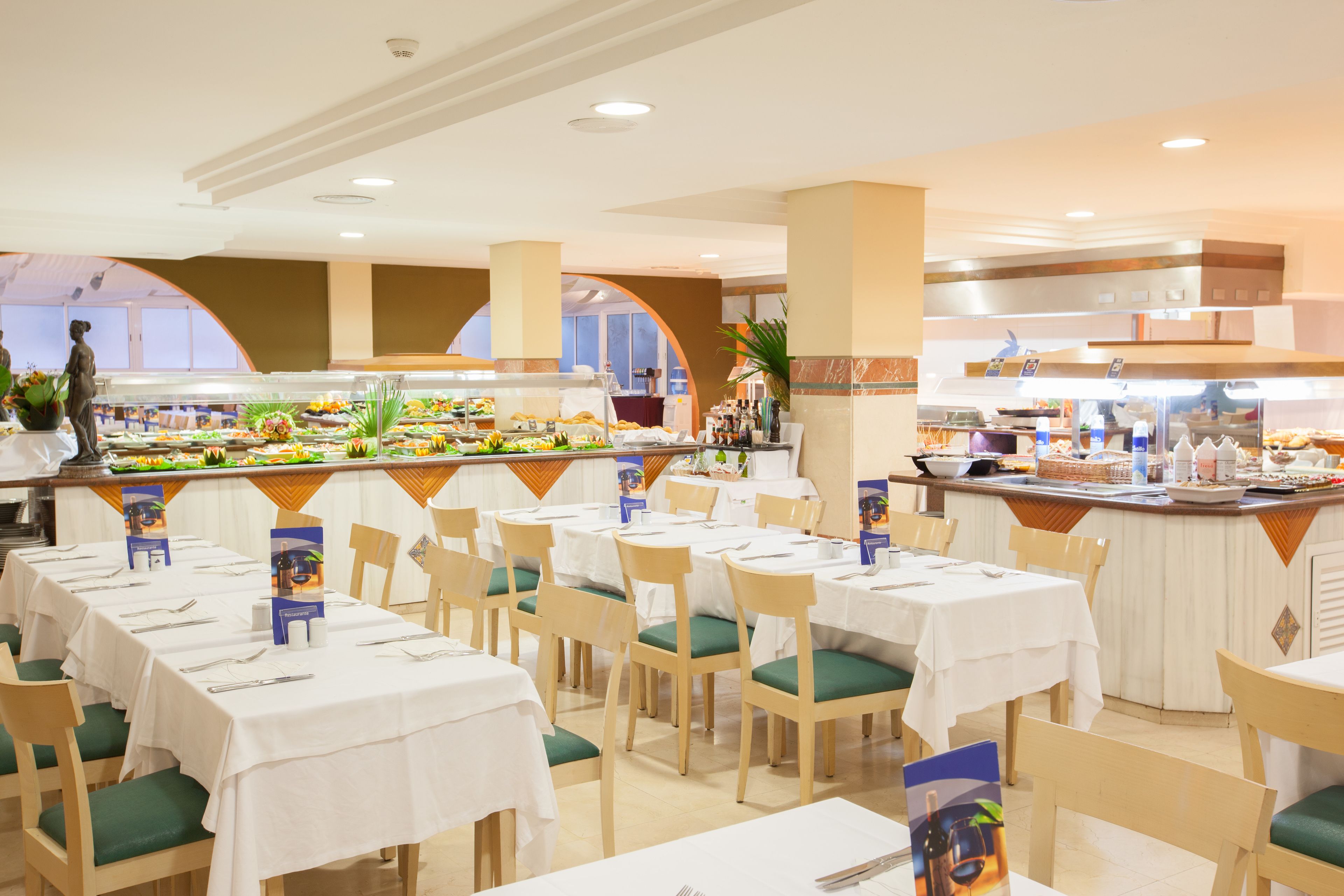 Un área de buffet con varias mesas y sillas, un mostrador central de comida y una cocina al fondo.