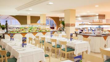 Un área de buffet con varias mesas y sillas, un mostrador central de comida y una cocina al fondo.