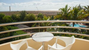 Un balcón con dos sillas blancas y una mesa redonda pequeña, con vista a una playa y una piscina.
