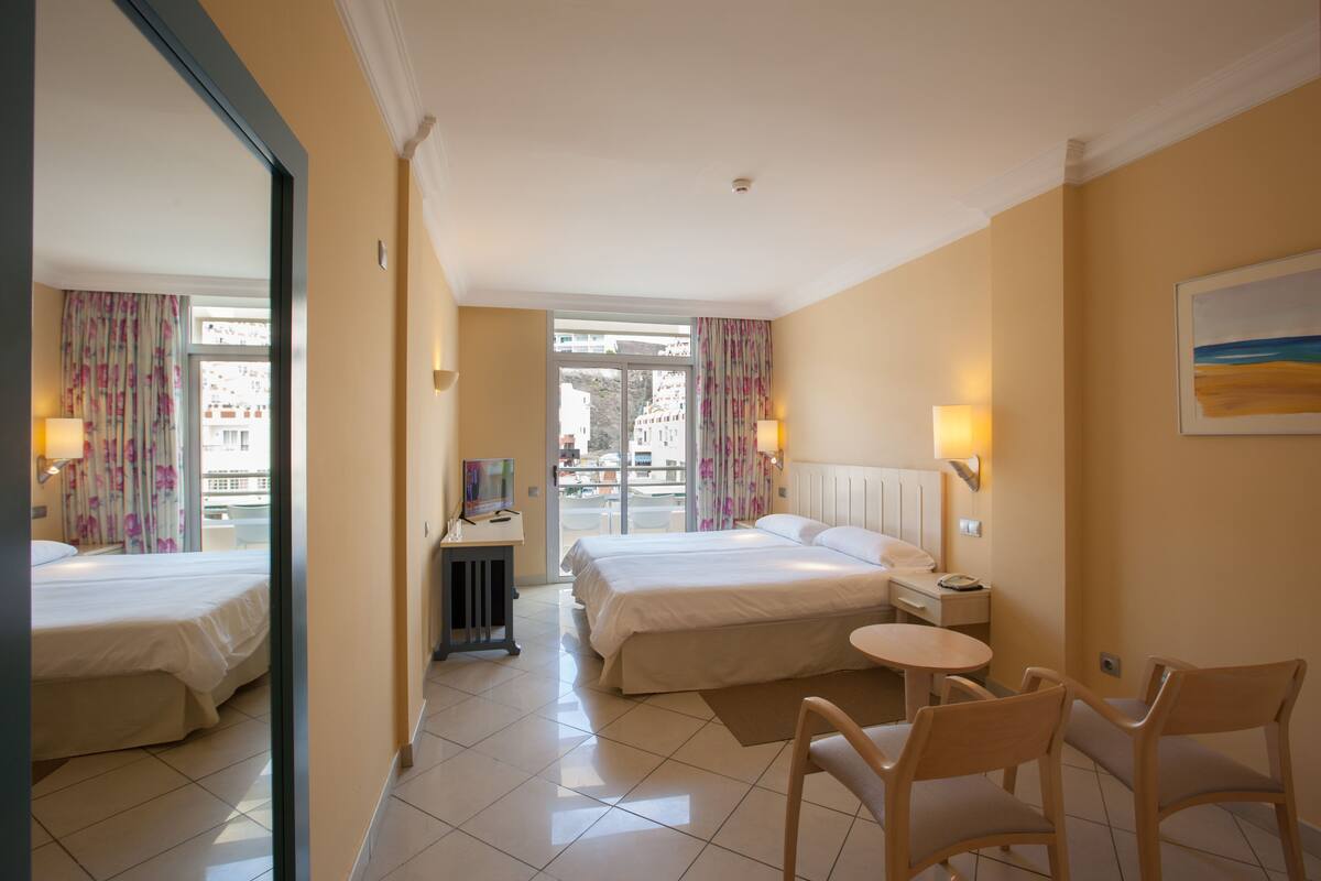 A hotel room with two beds, a desk with a chair, a small table, and a view of the outside.