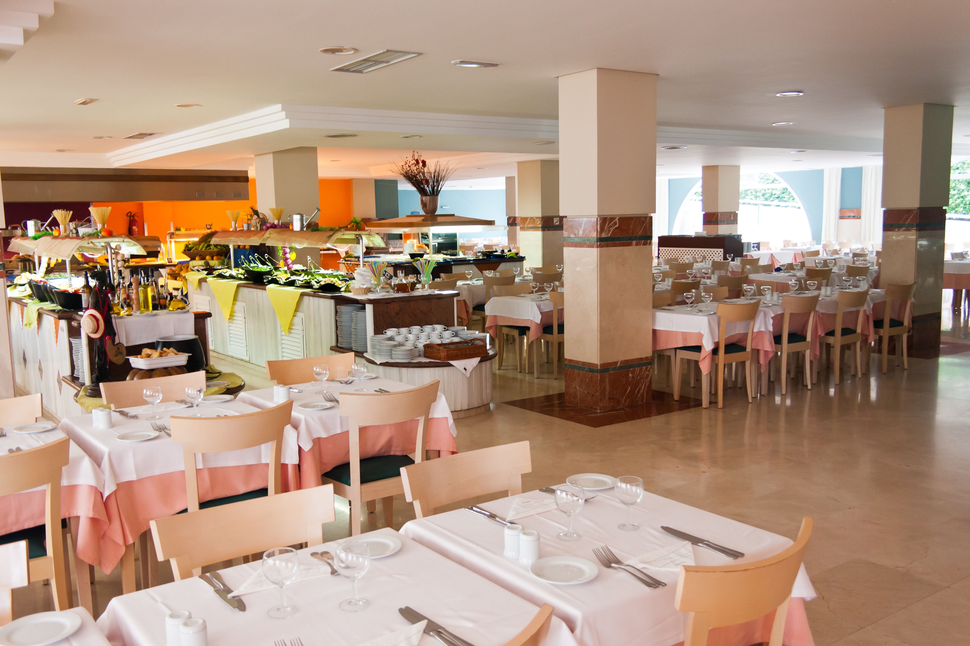 Une salle à manger d’hôtel avec de nombreuses tables dressées pour les clients, un buffet et une cuisine en arrière-plan.